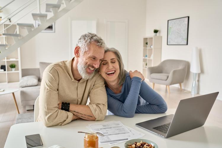 Couple sitting at a table, looking at a laptop, smiling or laughing.
